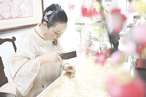 ◎ 一片薄如蟬翼的銅箔,一把帶圓珠頭的鋼鑿,在木槌的敲打下,銅箔片上出現了凹陷的珠點。不斷敲打下,以點成線、以線成面、勾勒出一幅金碧輝煌的畫面......近日,在廣東佛山饒寶蓮金銅鑿剪紙藝術館,饒寶蓮正在為當地博物館趕製一幅包含佛山十五項非遺主題的銅鑿剪紙。圖為八月十九日,廣東省佛山市,饒寶蓮使用木槌和帶圓珠頭的鋼鑿製作銅鑿剪紙。(中新社記者 陳驥旻 攝)