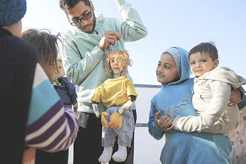 ◎ 巴勒斯坦志願者近期在加沙地帶多地開展各類志願活動,幫助兒童減輕因持續戰亂帶來的心理創傷。圖為2月19日,在加沙地帶南部城市拉法的一處臨時營地,一名巴勒斯坦志願者用自製的提線木偶為孩子們表演。(新華社發 里澤克·阿卜杜勒賈瓦德 攝)