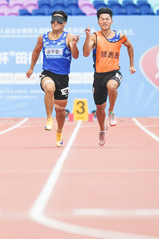 ◎ 十二月十一日,遼寧隊選手趙平安(左)和領跑員鍾才在男子一百米T11級決賽中。最終,他獲得冠軍。第十二屆全國殘疾人運動會暨第九屆特殊奧林匹克運動會田徑男子一百米T11級決賽在廣州舉行。(新華社記者 劉夢琪 攝)