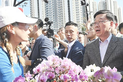 ◎ 二月十六日,農曆乙巳蛇年除夕,香港特區行政長官李家超前往觀塘年宵市場選購年花,並與市民及攤主交談。圖為李家超(前排右一)與攤主交談並購買年花。(中新社記者 李志華 攝)